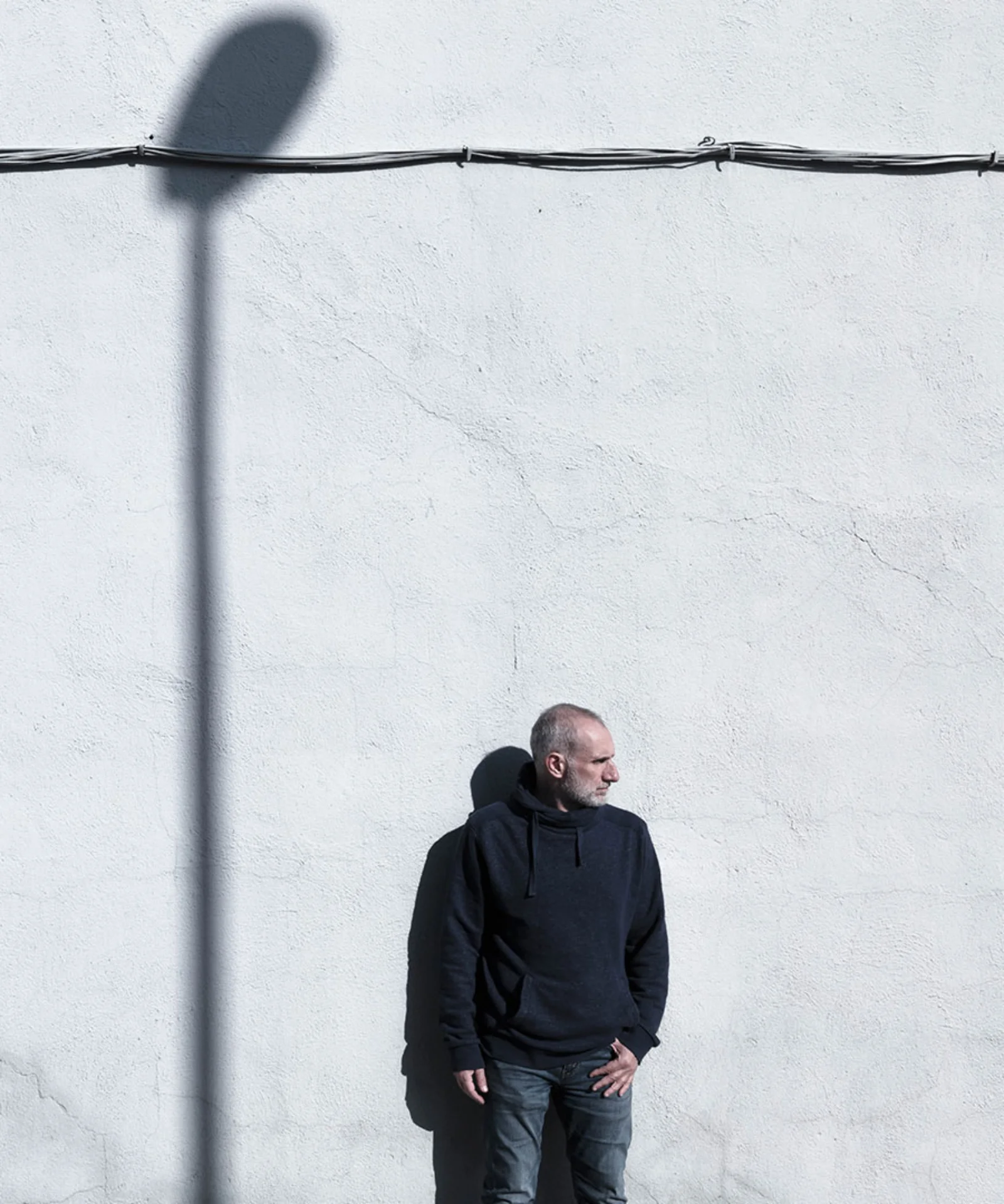 Man standing by minimalist wall with strong shadows Man in dark clothing standing against a textured white wall with geometric shadows, evoking contrast and contemplation.