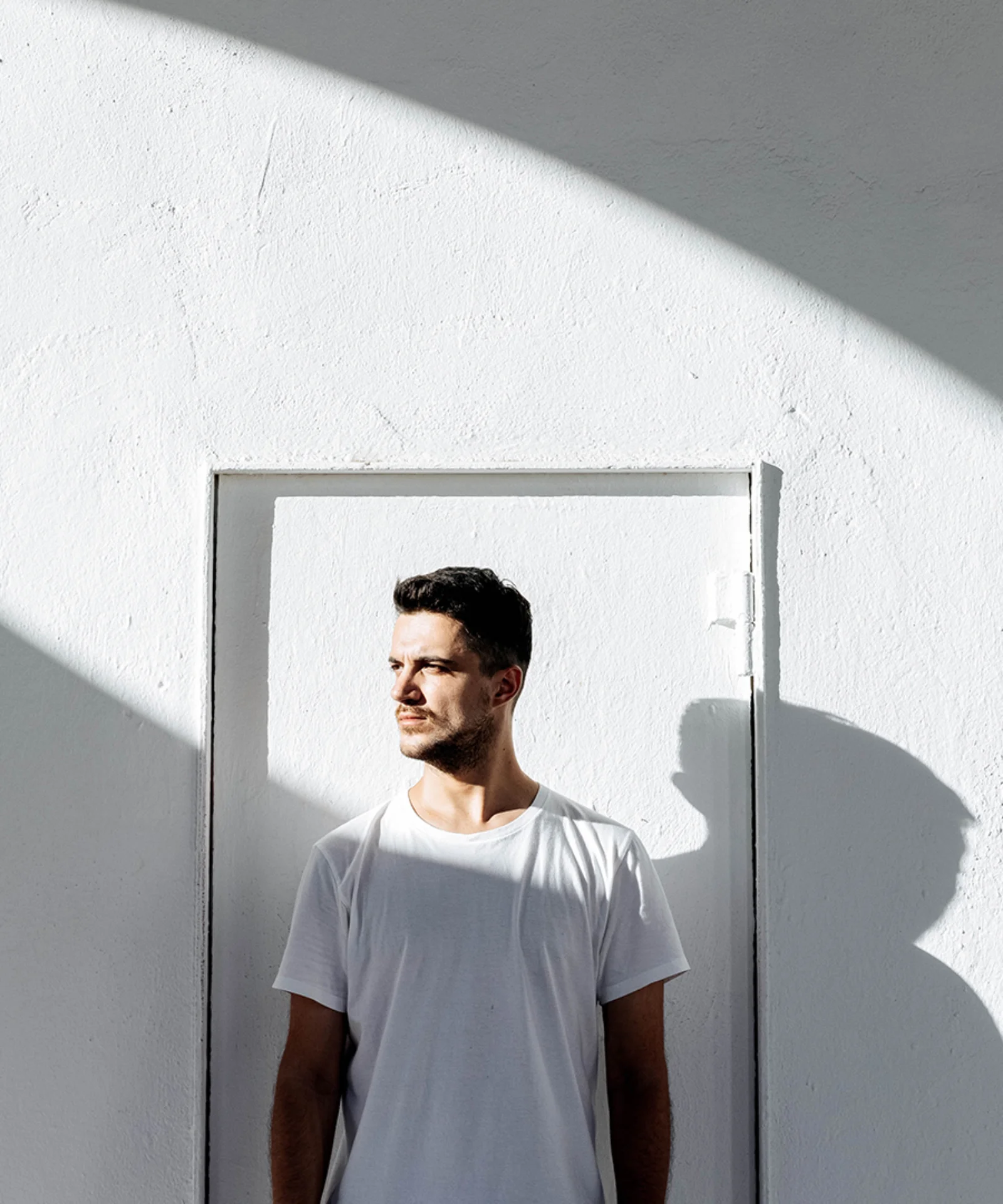 Person standing against a minimalist white wall with sunlight casting a circular shadow, creating a striking modern composition.