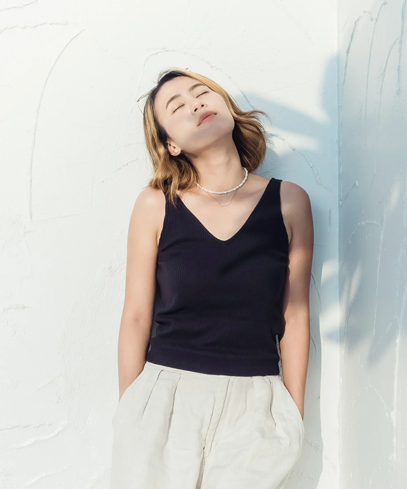 Person standing against a white textured wall with eyes closed, relaxed and bathed in natural sunlight.