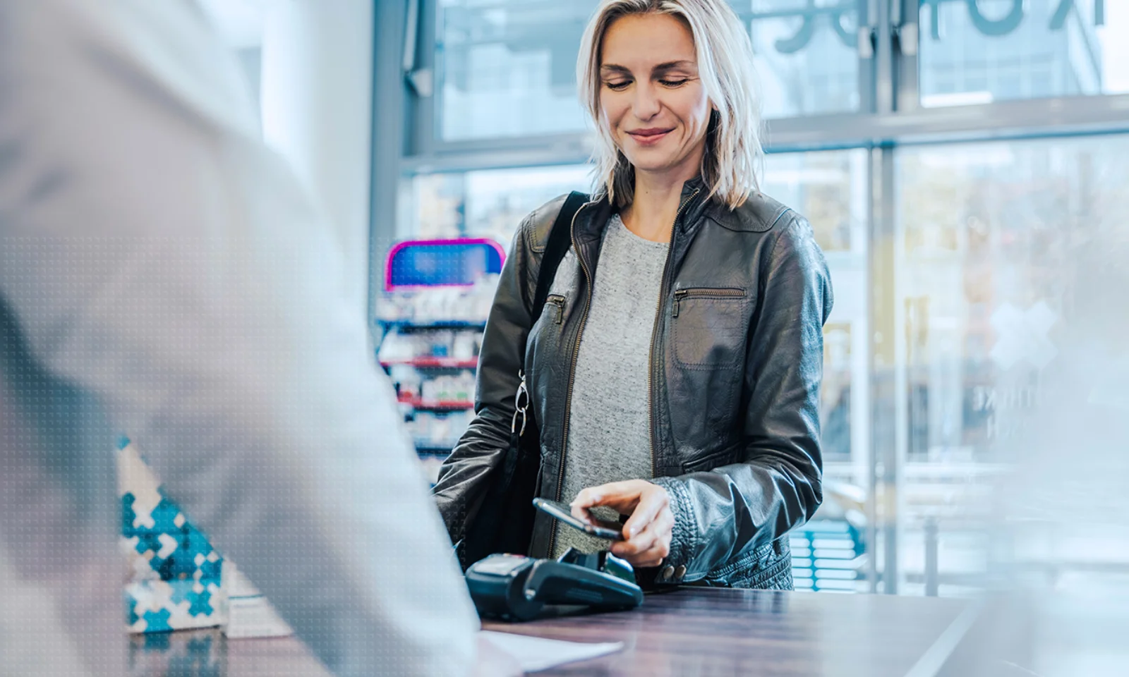 GFT US Blog ICYMI Moneylive 2024 - 1 A woman in a leather jacket makes a contactless payment with her smartphone at a retail store counter, demonstrating modern digital payment technology.