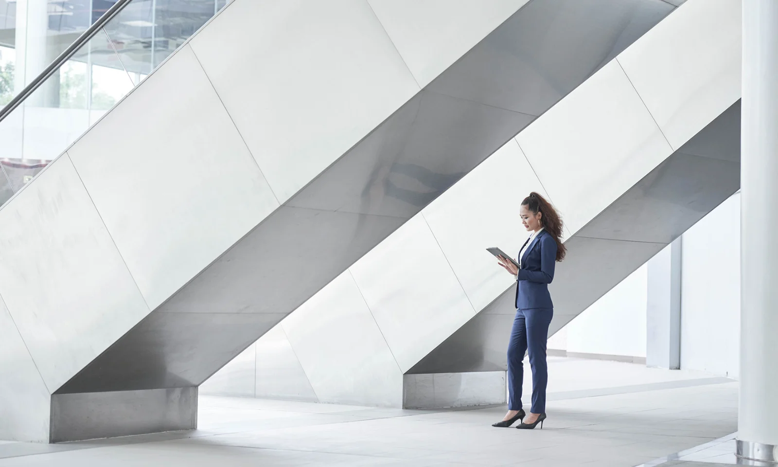 Woman in a blue suit standing in a bright, modern building using a tablet, symbolizing digital innovation and professional efficiency.