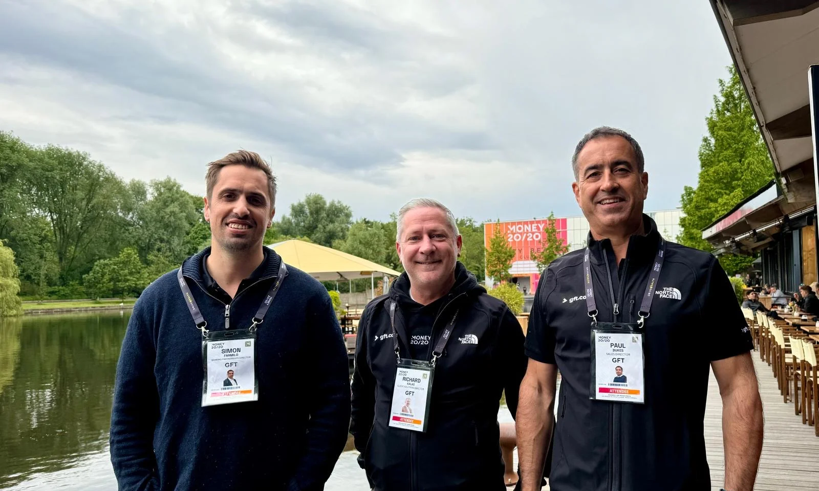 Three GFT representatives standing outdoors at the Money20/20 event, wearing event badges and GFT-branded attire, with a lake and trees in the background.