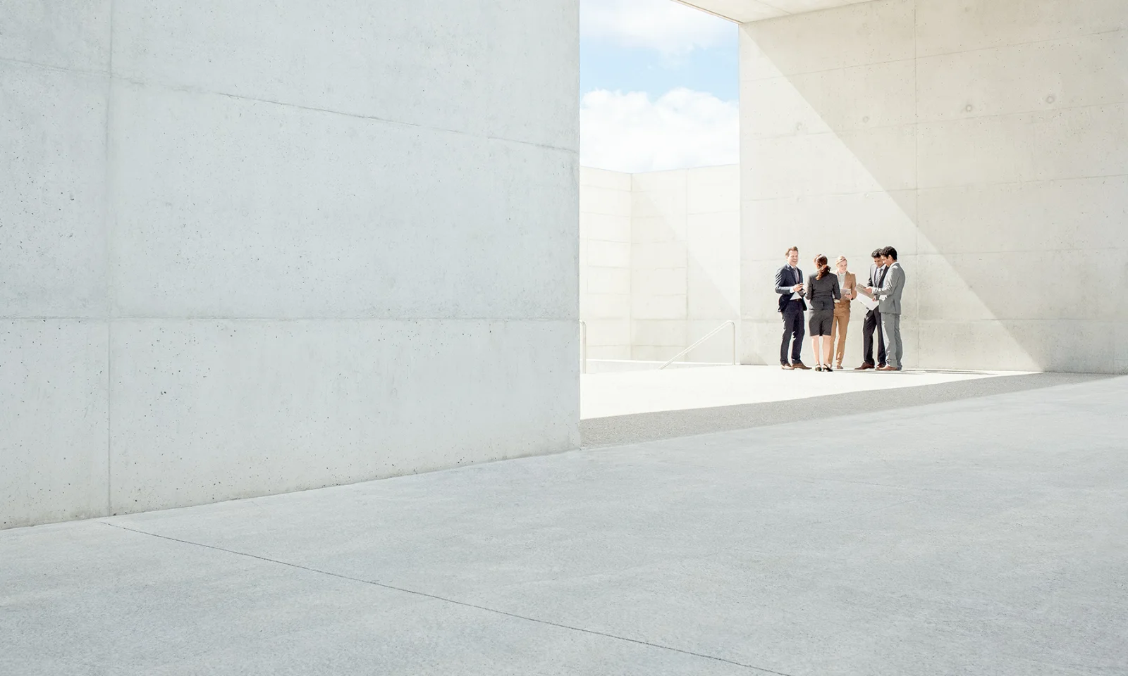 GFT Blog: On The FedNow Service: What To Expect From The First Version Group of business professionals standing together in a bright, open, minimalist concrete space, collaborating and reviewing documents under natural light.