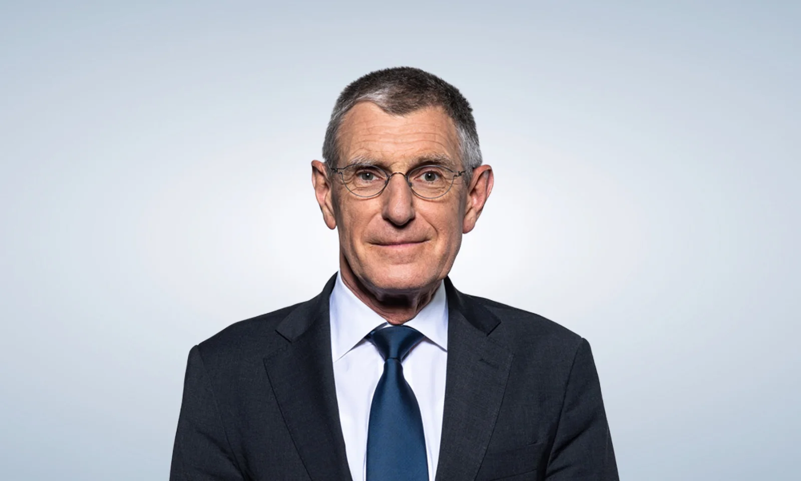 Executive Board Member – Corporate Governance Professional portrait of a smiling business executive wearing glasses, a dark suit, blue shirt, and green patterned tie against a light gradient background.