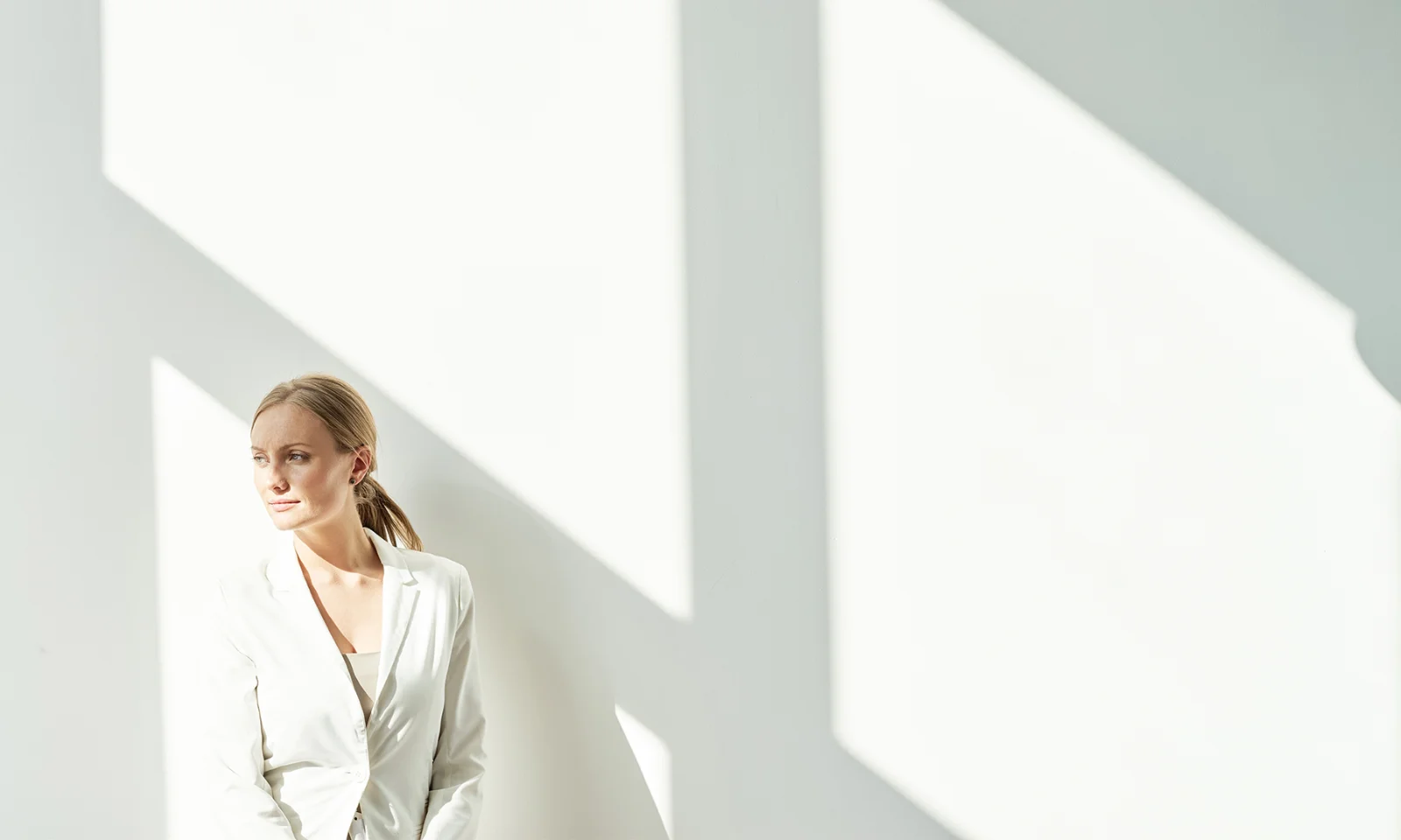 GFT Image - Mood - 04 Person in a white blazer standing in bright natural light against a minimalist white wall, looking thoughtfully into the distance.