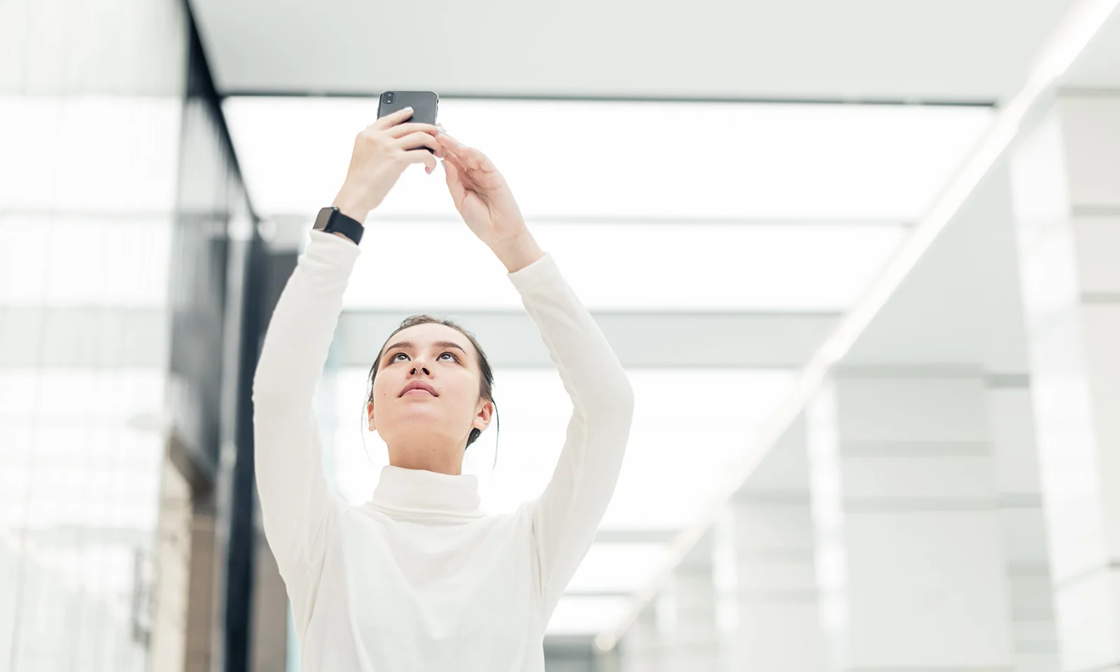 Woman Using Smartphone in Modern Office Environment Person holding a smartphone upwards in a bright, modern office setting, symbolizing connectivity, innovation, and digital transformation.