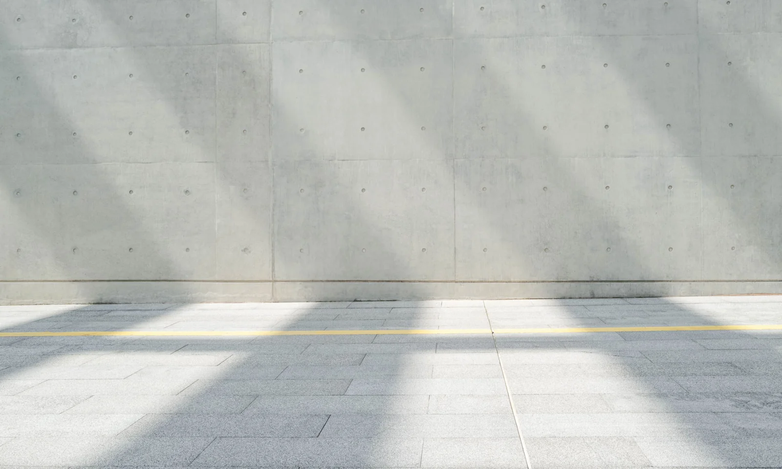 Modern Concrete Wall with Light and Shadow Patterns Smooth concrete wall with visible panel joints and soft vertical shadows falling across a tiled pavement with a yellow line.