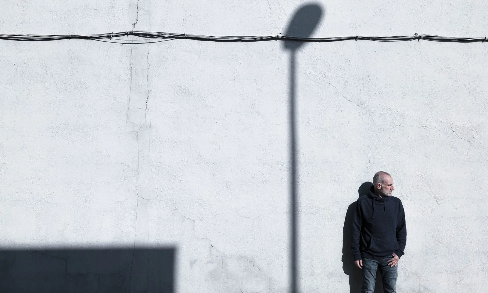 Man standing by minimalist wall with strong shadows Man in dark clothing standing against a textured white wall with geometric shadows, evoking contrast and contemplation.