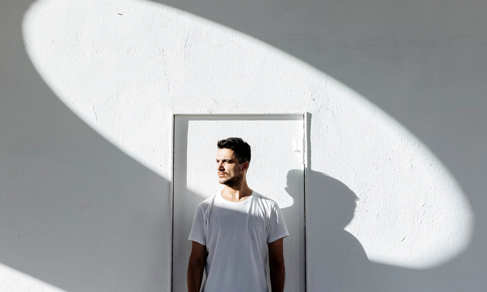 GFT Blog - Segurança cibernética é vital na transformação digital Person standing against a minimalist white wall with sunlight casting a circular shadow, creating a striking modern composition.