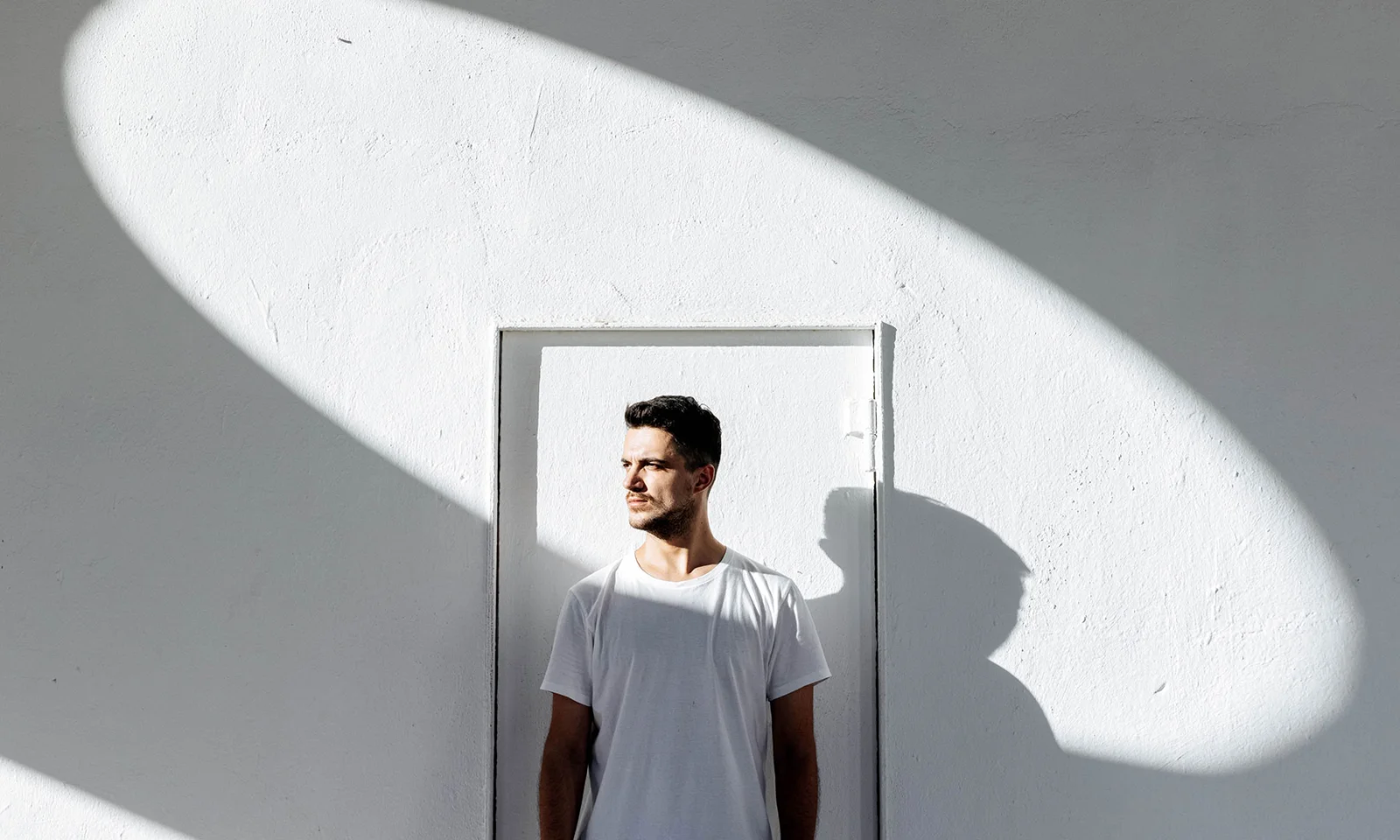 Man standing in sunlight against minimalist white wall Man in white shirt standing in bright sunlight, framed by geometric shadows on a white wall, symbolizing clarity and innovation.