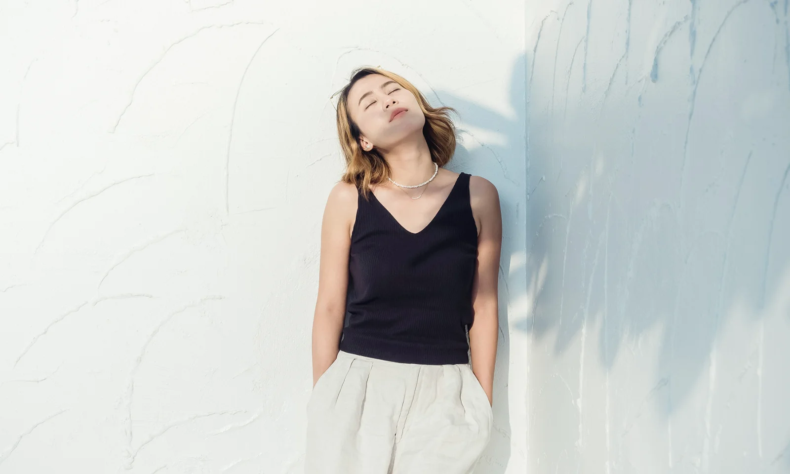 GFT Blog - O novo CEO em 2025: Algoritmos, cultura digital e o peso da reinvencao pessoal Person standing against a white textured wall with eyes closed, relaxed and bathed in natural sunlight.