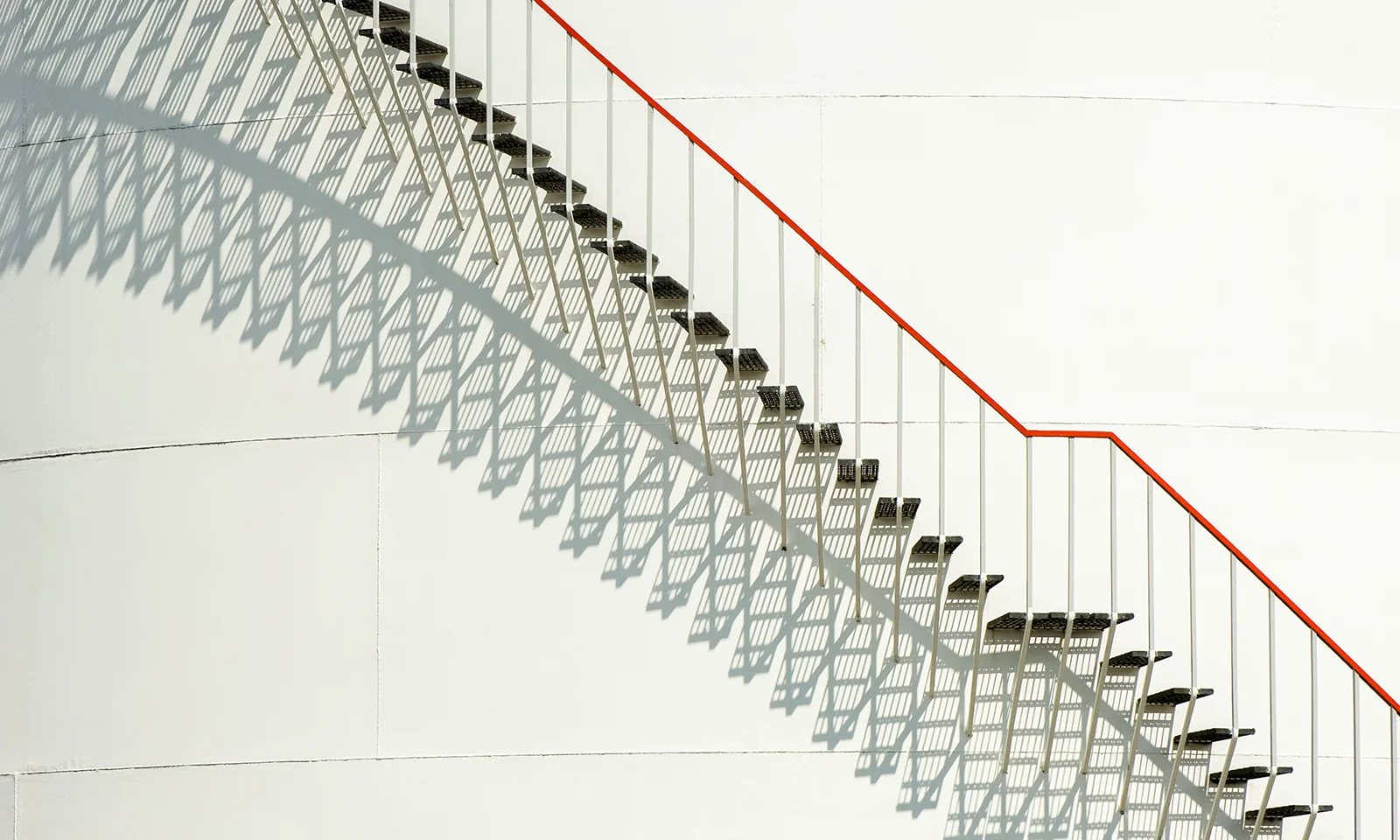 Modern Industrial Staircase with Geometric Shadows Metal staircase with red handrail casting geometric shadows on a white industrial wall in natural light