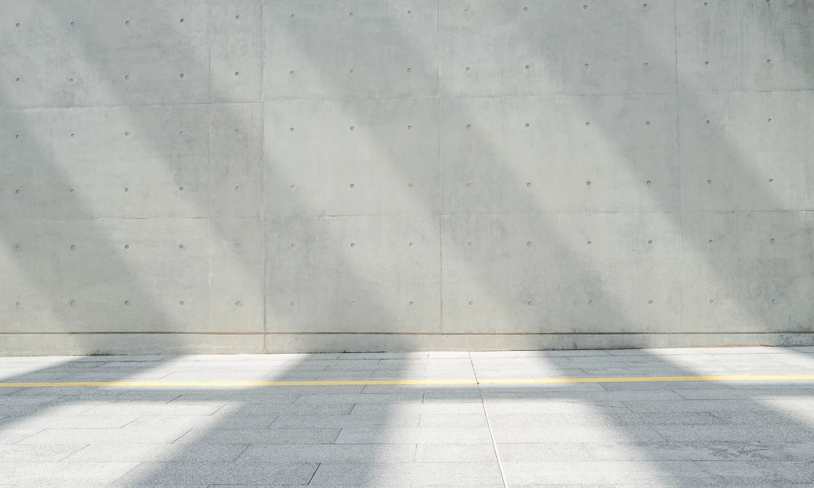 Concrete Wall with Linear Shadows and Yellow Accent Modern concrete wall with evenly cast shadows and a single yellow line across a tiled pavement, creating a minimalist architectural composition.
