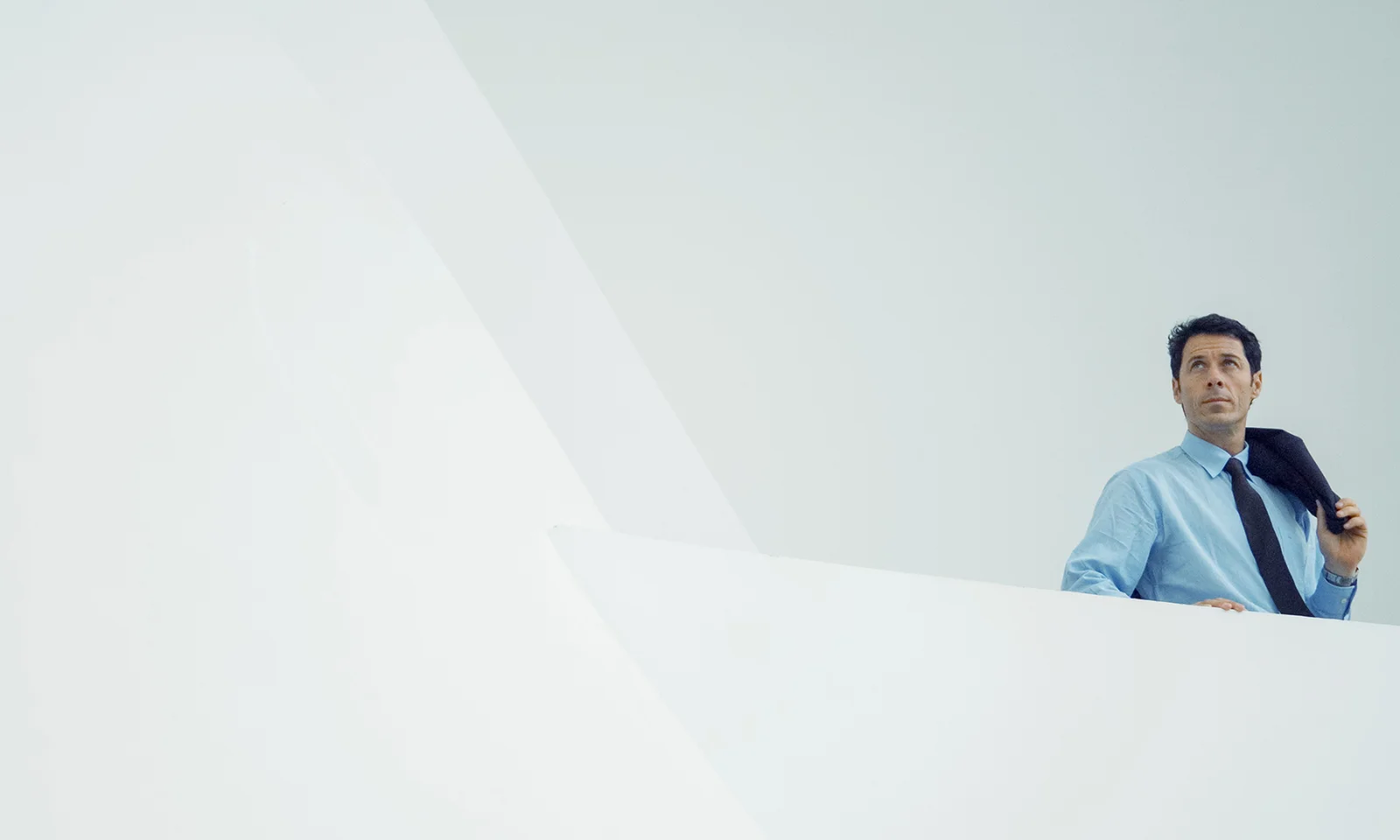 GFT Blog - Como a IA está redefinindo o jogo no setor de Telecomunicações Man in business attire holding a jacket over his shoulder, standing in a minimalist white architectural setting, looking upward with a confident expression.