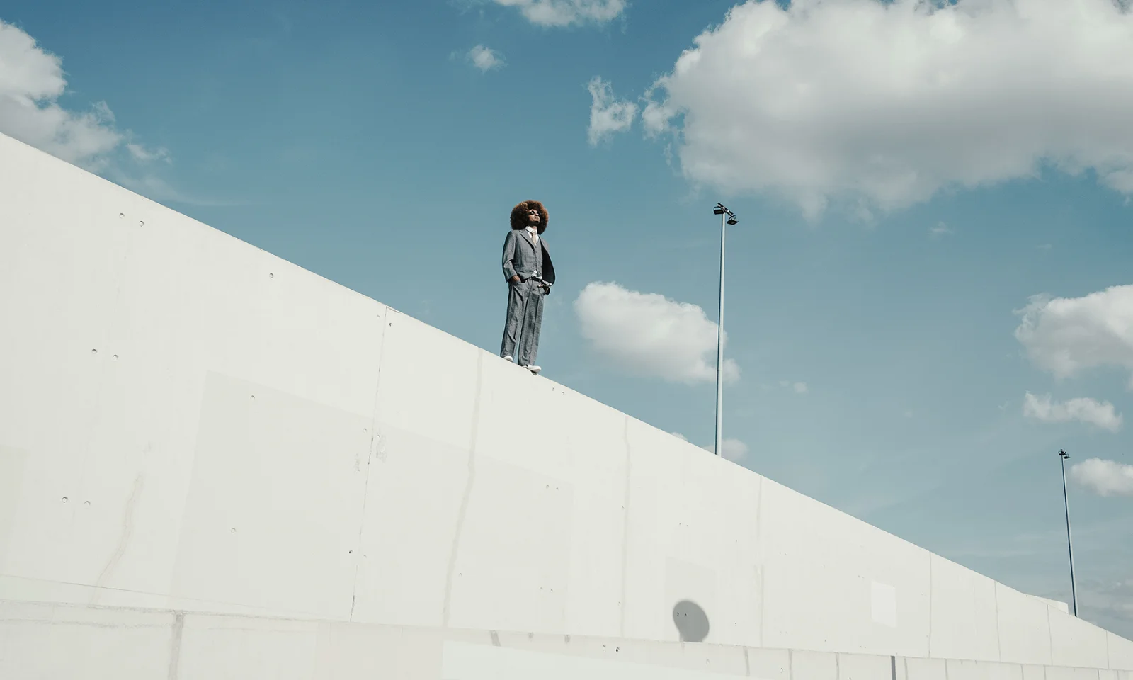 Confident Professional Standing on Modern Architectural Structure Under Blue Sky Person in a suit standing atop a minimalist concrete wall with a bright blue sky and clouds in the background