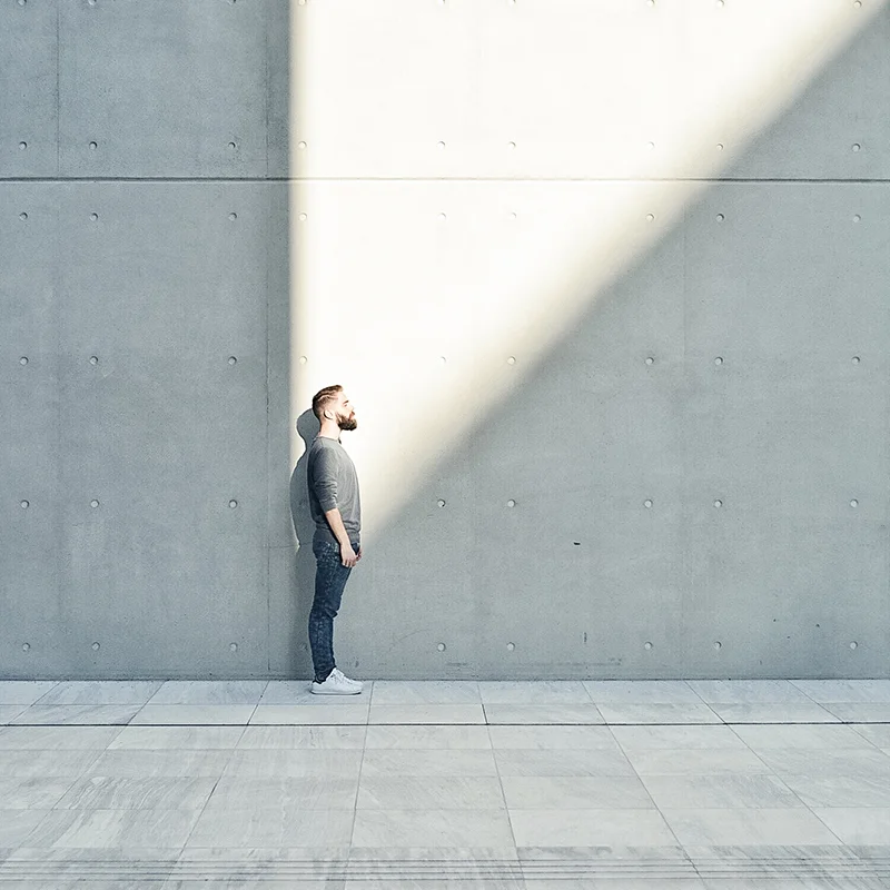 Innovation Mindset – Vision and Perspective Person standing against a minimalist concrete wall, looking up toward a bright beam of sunlight, symbolizing inspiration and forward thinking.