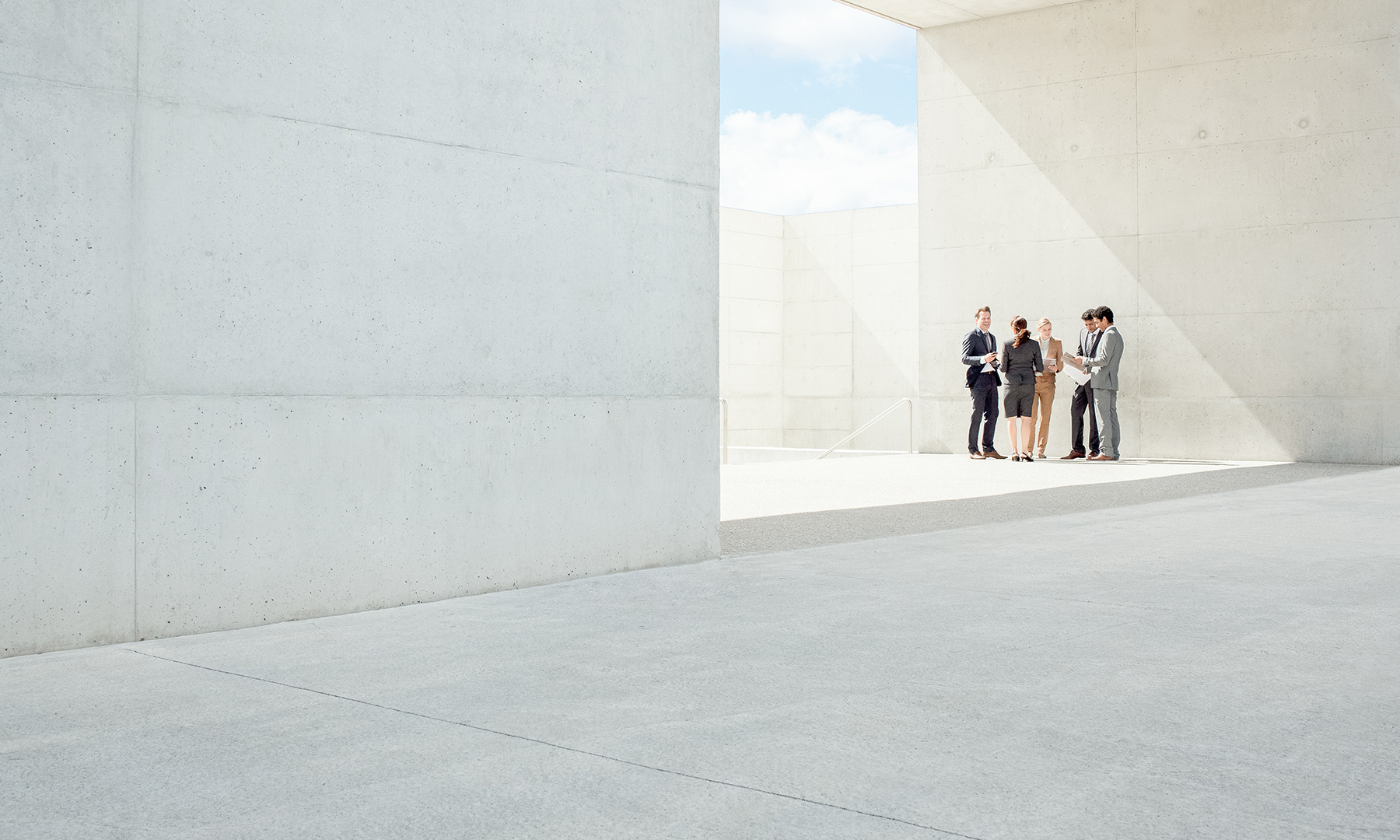Group of business professionals standing together in a bright, open, minimalist concrete space, collaborating and reviewing documents under natural light.