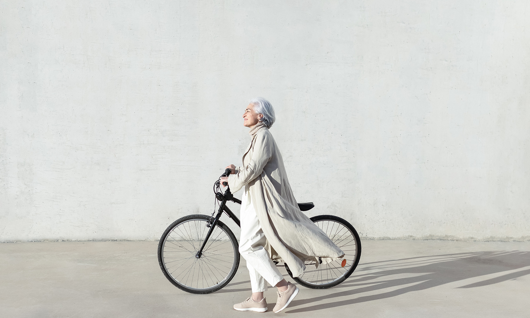 GFT Success Story: A Groundbreaking Cloud Native Architecture for Credit Risk HPC-Processing Woman with white hair walking beside a bicycle in front of a light gray wall, dressed in neutral tones under soft sunlight.