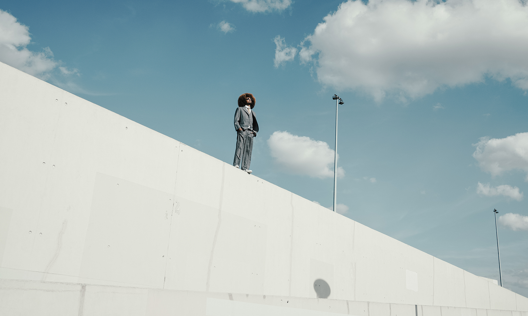 Person in a suit standing confidently on a high white wall under a bright blue sky with clouds, symbolizing vision and leadership.