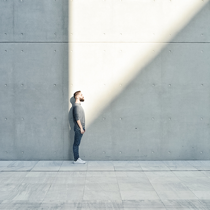 Innovation Mindset – Vision and Perspective Person standing against a minimalist concrete wall, looking up toward a bright beam of sunlight, symbolizing inspiration and forward thinking.
