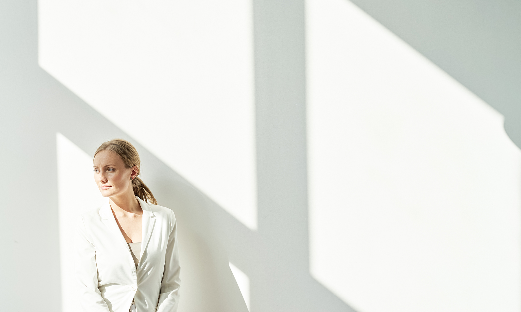 Person in a white blazer standing in bright natural light against a minimalist white wall, looking thoughtfully into the distance.