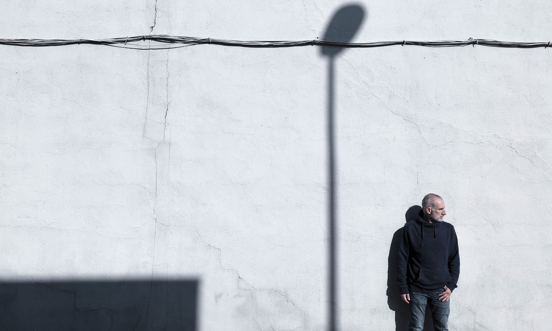 Man in dark clothing standing against a textured white wall with geometric shadows, evoking contrast and contemplation.