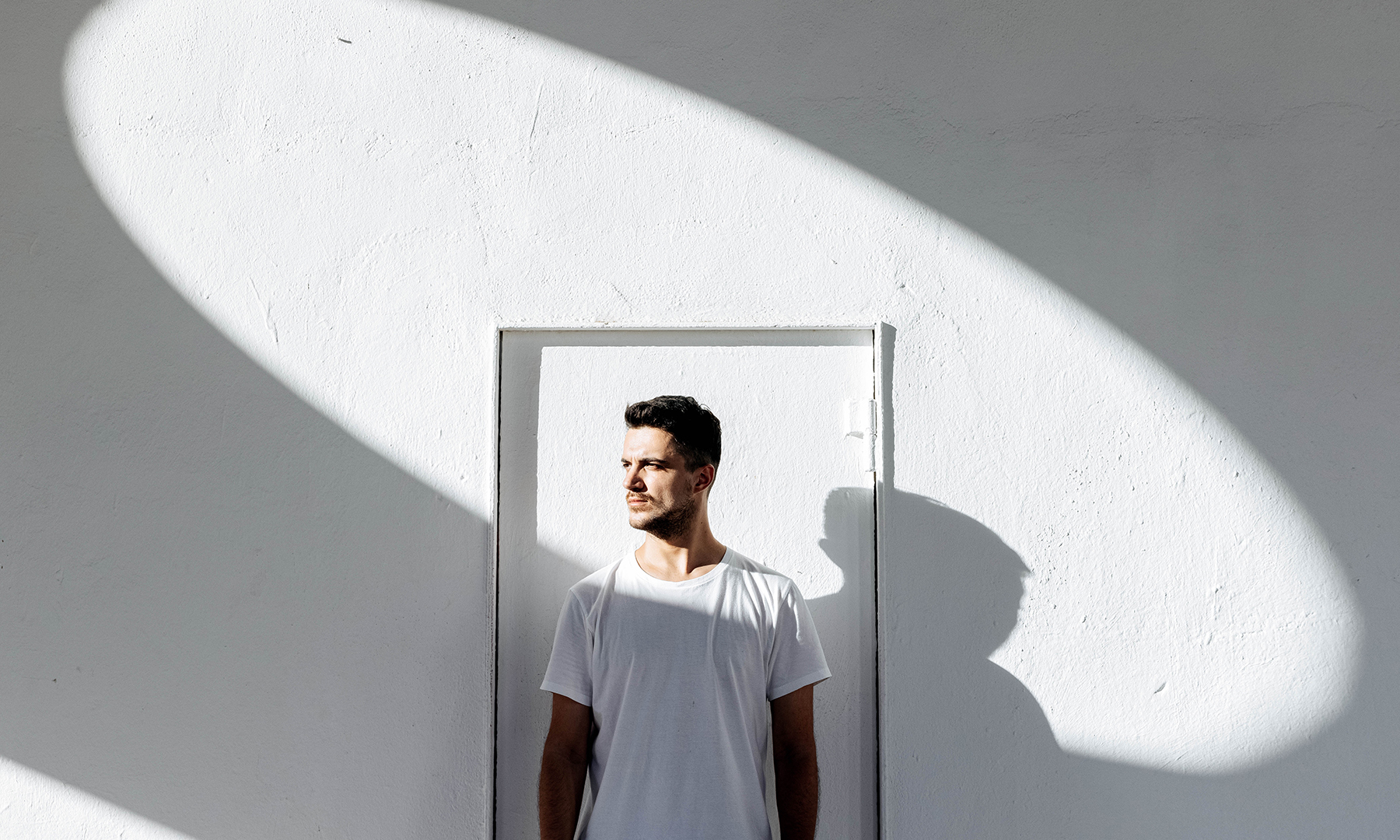 Person standing against a minimalist white wall with sunlight casting a circular shadow, creating a striking modern composition.