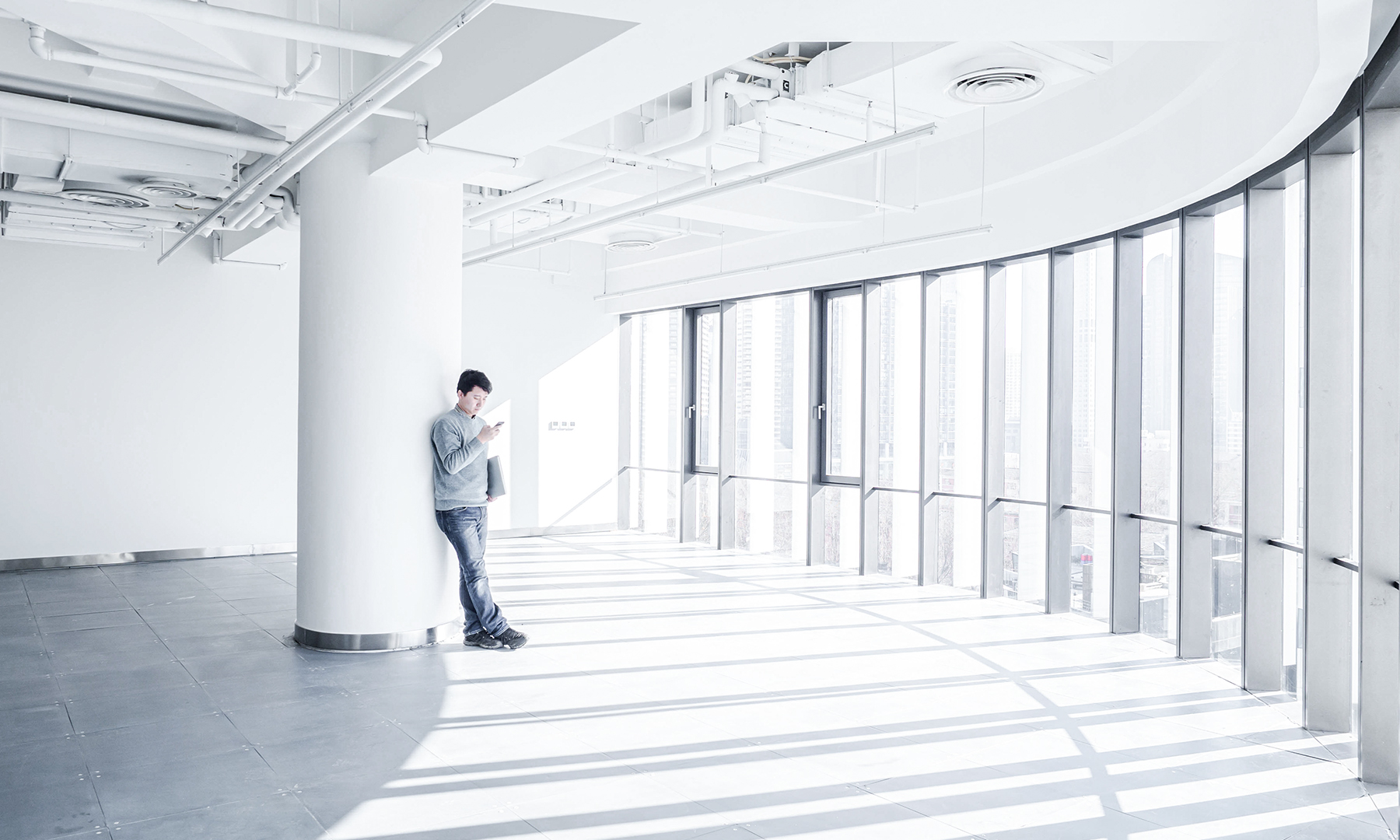 Professional Reading in Bright Modern Office Space Man leaning against a white column in a spacious, sunlit office with large windows and minimalist industrial design.