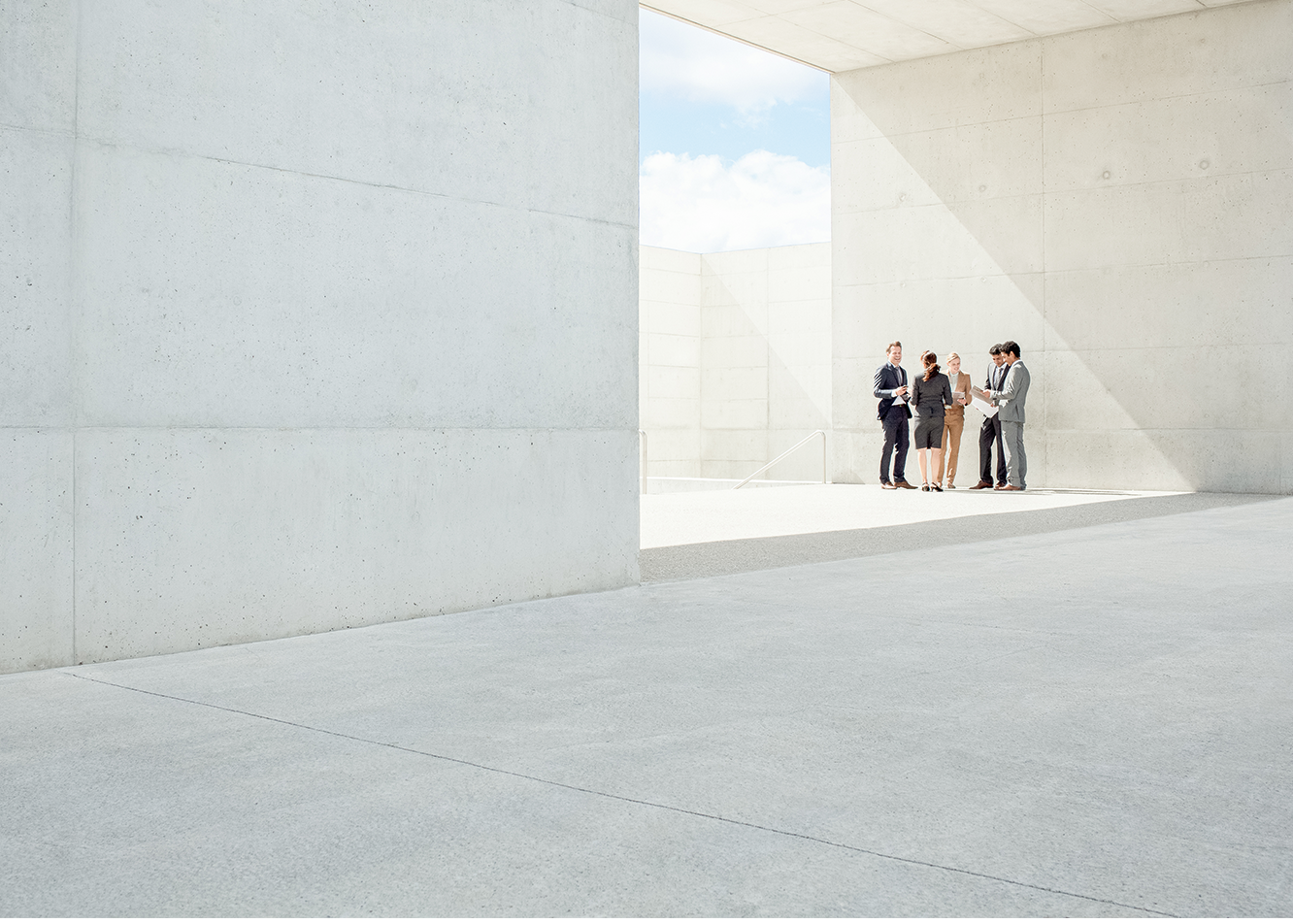 Eine Gruppe von Leuten steht vor einer grauen Wand. Sonne scheint von oben auf die Personen