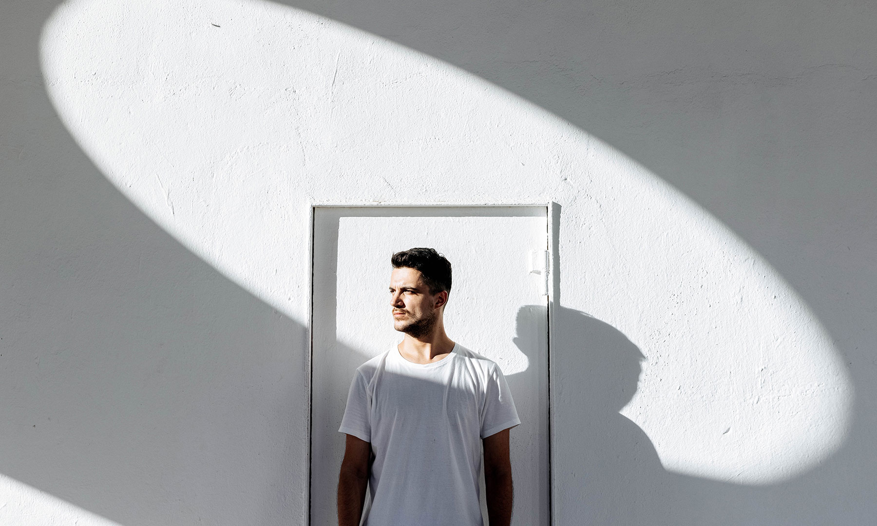 Man Standing in Minimalist Space with Dramatic Light and Shadow Man in a white t-shirt standing against a bright white wall, illuminated by a bold oval shadow pattern cast by sunlight.