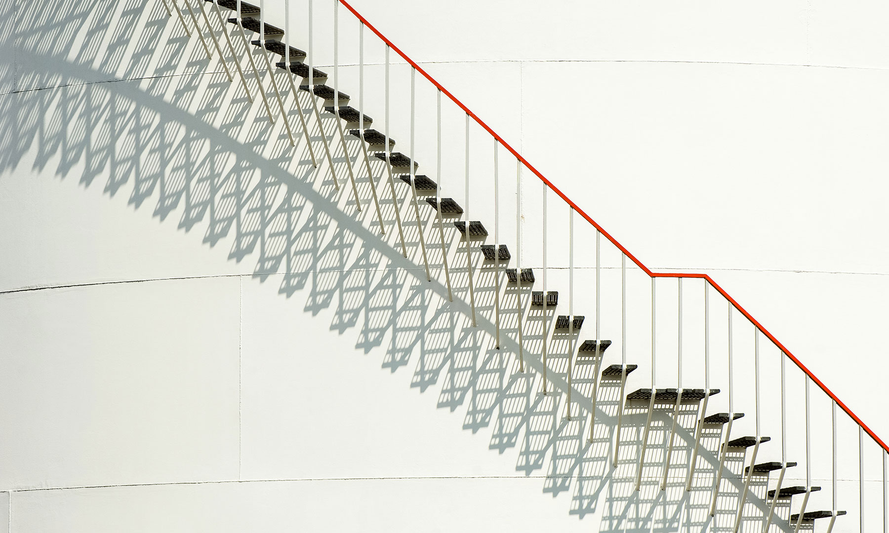 GFT Success Story: Healthcare Manufacturer Transforms HR Minimalist metal staircase with a red handrail casting geometric shadows on a clean white wall in natural light.