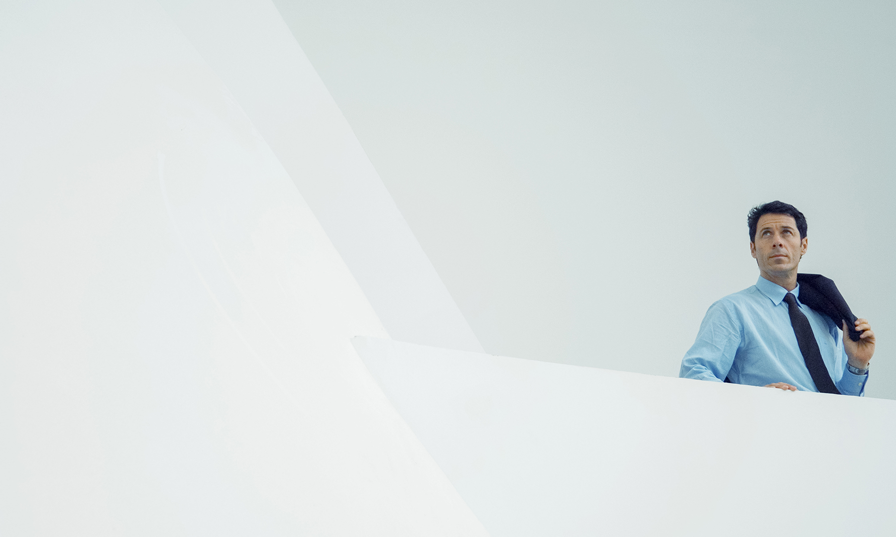 Man in business attire holding a jacket over his shoulder, standing in a minimalist white architectural setting, looking upward with a confident expression.